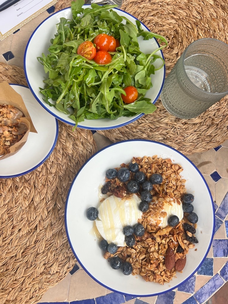 Casa Laguna Hotel and Spa breakfast spread on blue-rimmed enamel plates — granola with blueberries and yogurt, arugula salad with cherry tomatoes, served on a woven placemat on a Moroccan tile table