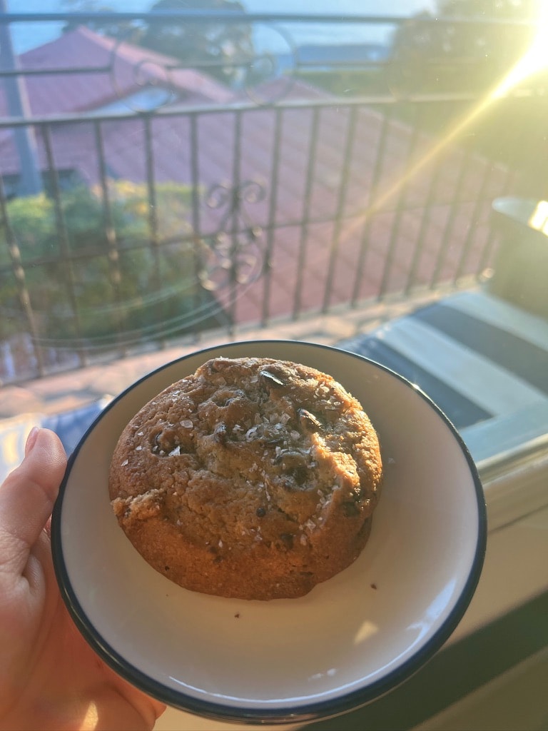 Casa Laguna Hotel and Spa sea-salt chocolate chip cookie on a blue-rimmed plate held up against a golden sunset view through the balcony railing, red tile rooftops and the Pacific visible below