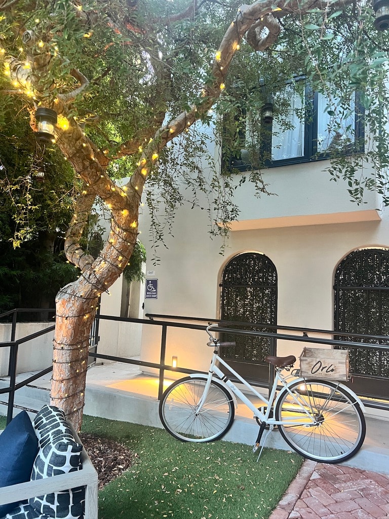 Orli La Jolla exterior at dusk with white bicycle, string-lit olive tree, and arched iron-screen facade
