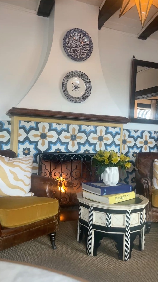 Casa Laguna Hotel and Spa interior sitting room with Moroccan-patterned fireplace tile surround, decorative blue-and-white plates on white plaster walls, leather armchairs, and a bone-inlay side table with yellow flowers