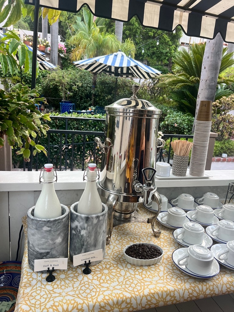 Casa Laguna Hotel and Spa outdoor morning coffee station with polished silver urn, Casa Laguna branded Half and Half and Almond Milk bottles in marble chillers, white cups and saucers, and striped umbrella overhead