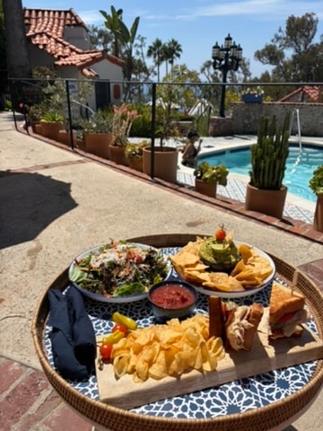 Casa Laguna Hotel and Spa poolside lunch tray with salad, guacamole, chips, and sandwich served on a round rattan tray beside the pool with gardens and terracotta pots in the background